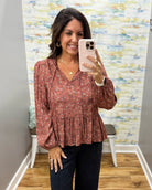 A woman takes a mirror selfie wearing a rust-colored long-sleeve blouse with a small floral print and dark pants in a fitting room.