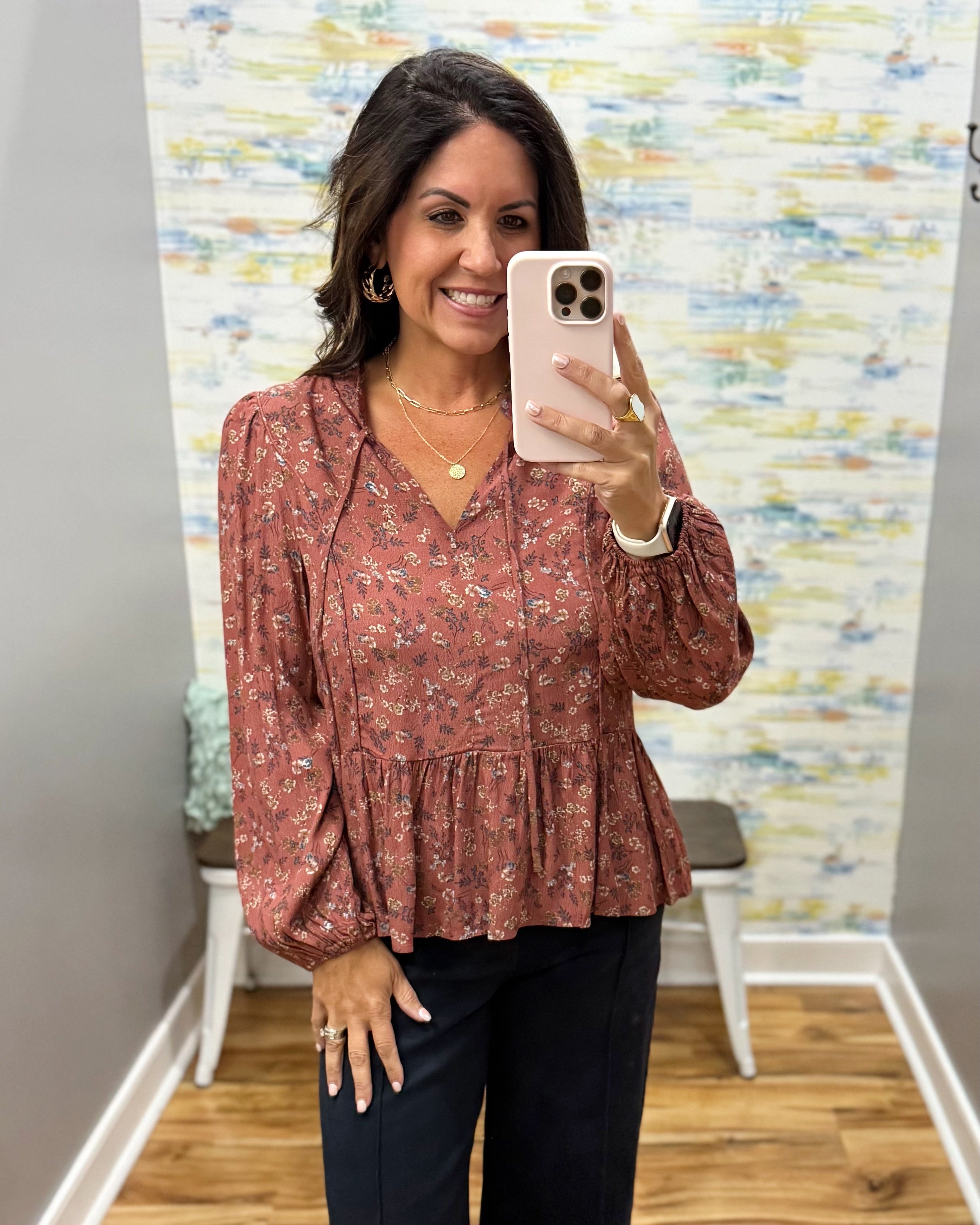 A woman takes a mirror selfie wearing a rust-colored long-sleeve blouse with a small floral print and dark pants in a fitting room.
