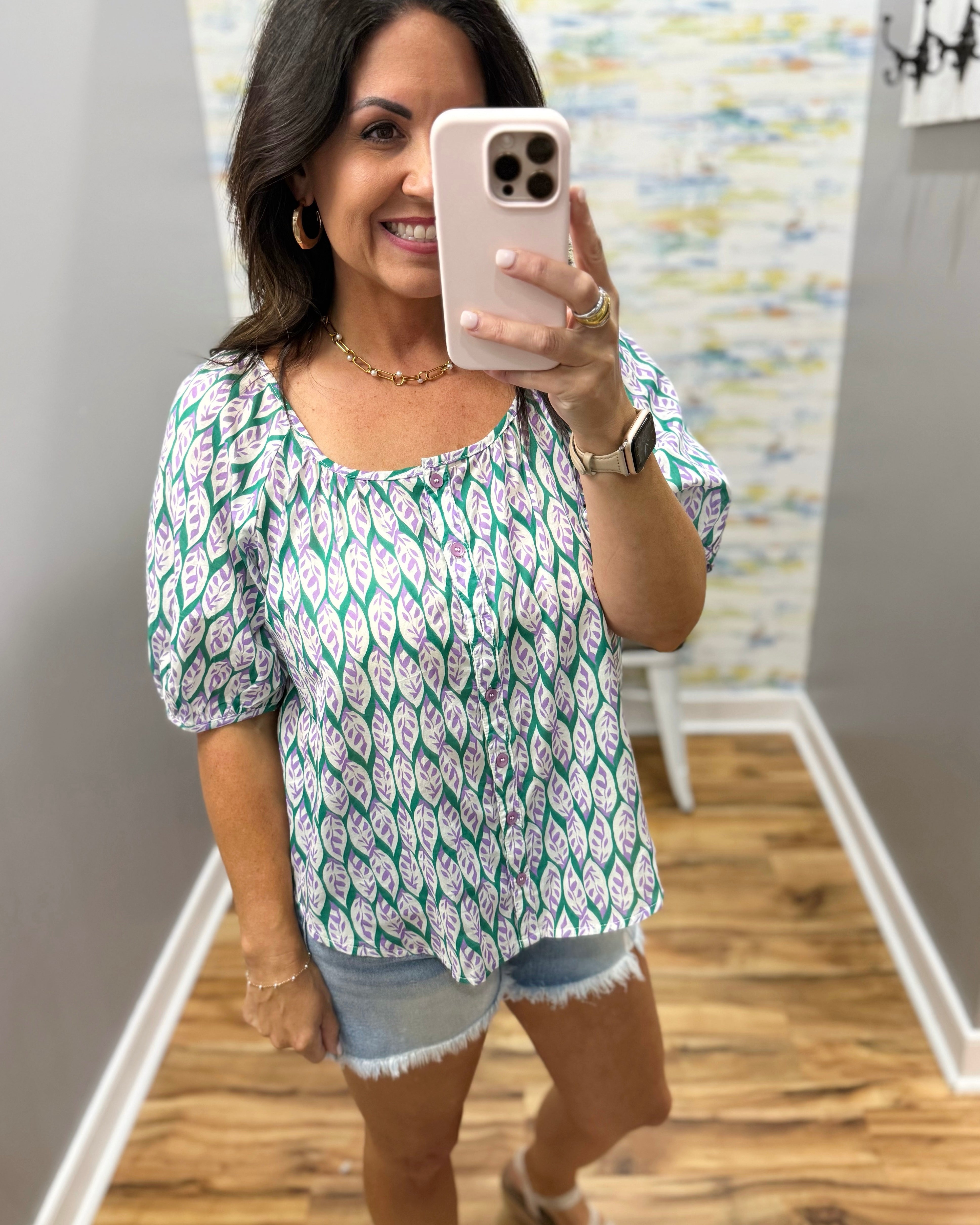 Woman taking a mirror selfie wearing a patterned top and denim shorts.
