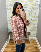 A woman takes a mirror selfie wearing a dusty rose and white plaid button-down shirt over a dark red top and blue jeans, standing in a room with a patterned wallpaper background.
