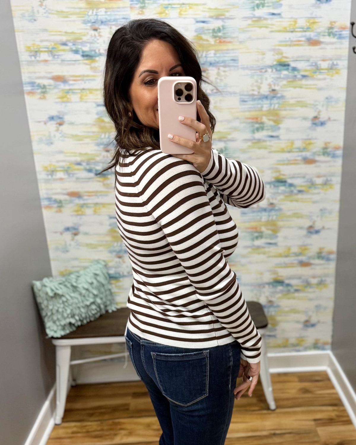 A woman taking a mirror selfie in a fitting room wearing a brown and white horizontally striped long-sleeve top and dark blue jeans.
