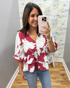 A woman takes a mirror selfie wearing a burgundy and white floral print blouse with blue jeans in an indoor boutique setting.