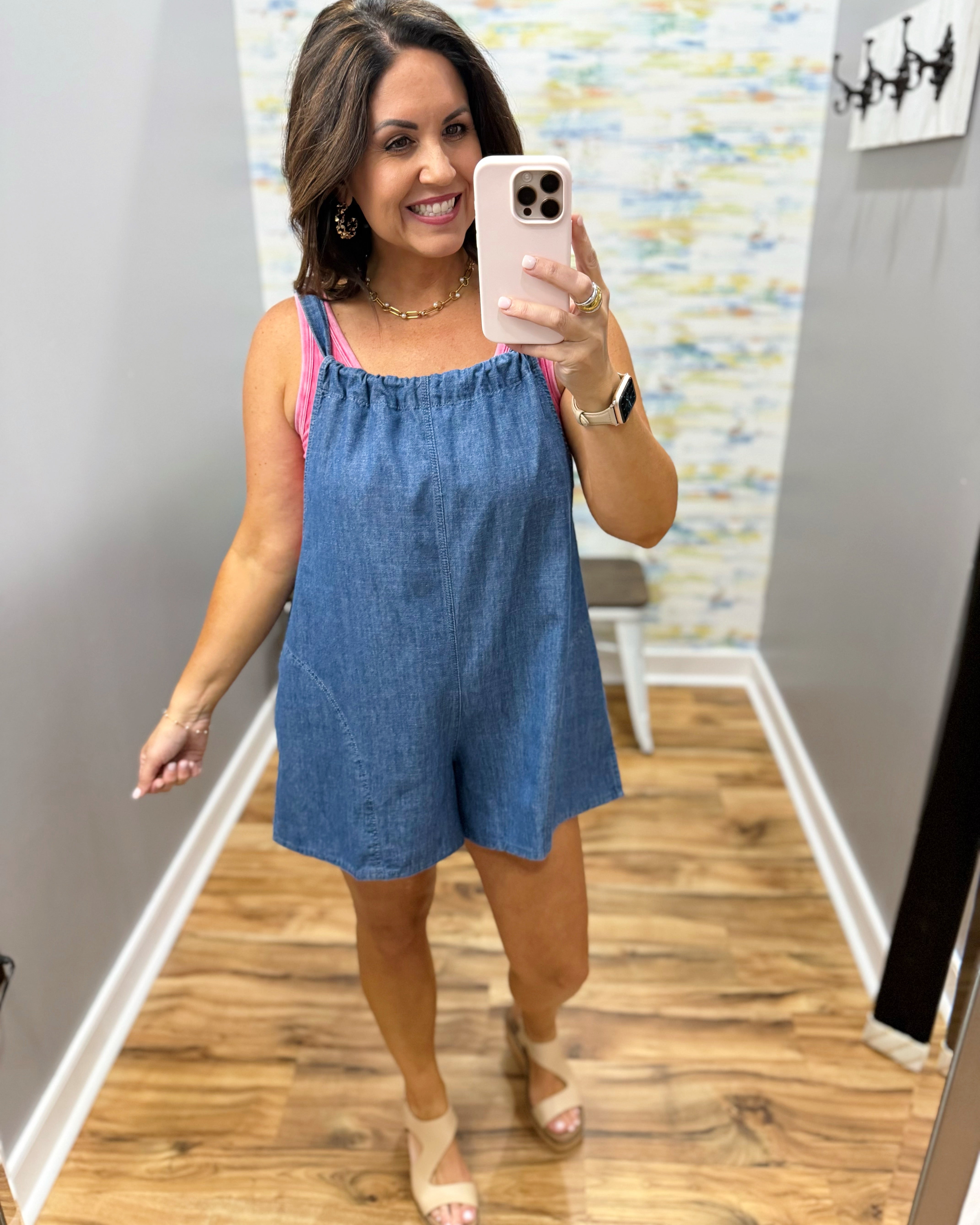 Woman taking a mirror selfie wearing a blue romper in a room with a colorful wall.