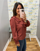 A woman takes a mirror selfie wearing a rust-colored long-sleeve blouse with ruffle details and blue jeans in a fitting room.