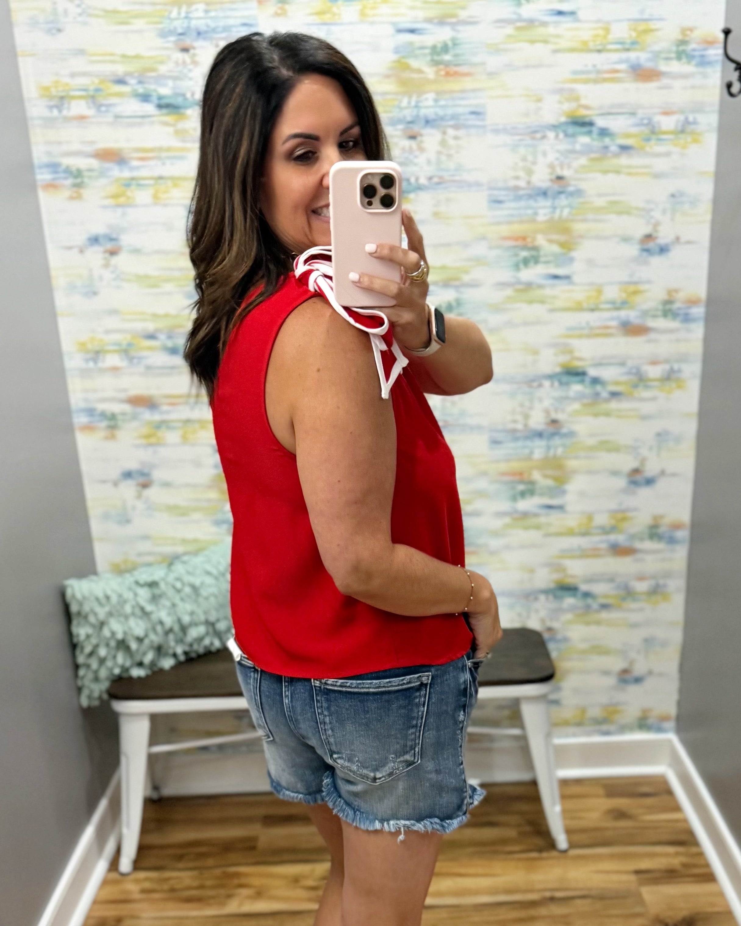 Woman taking a mirror selfie wearing a red sleeveless top and denim shorts in a room with colorful wall paper.