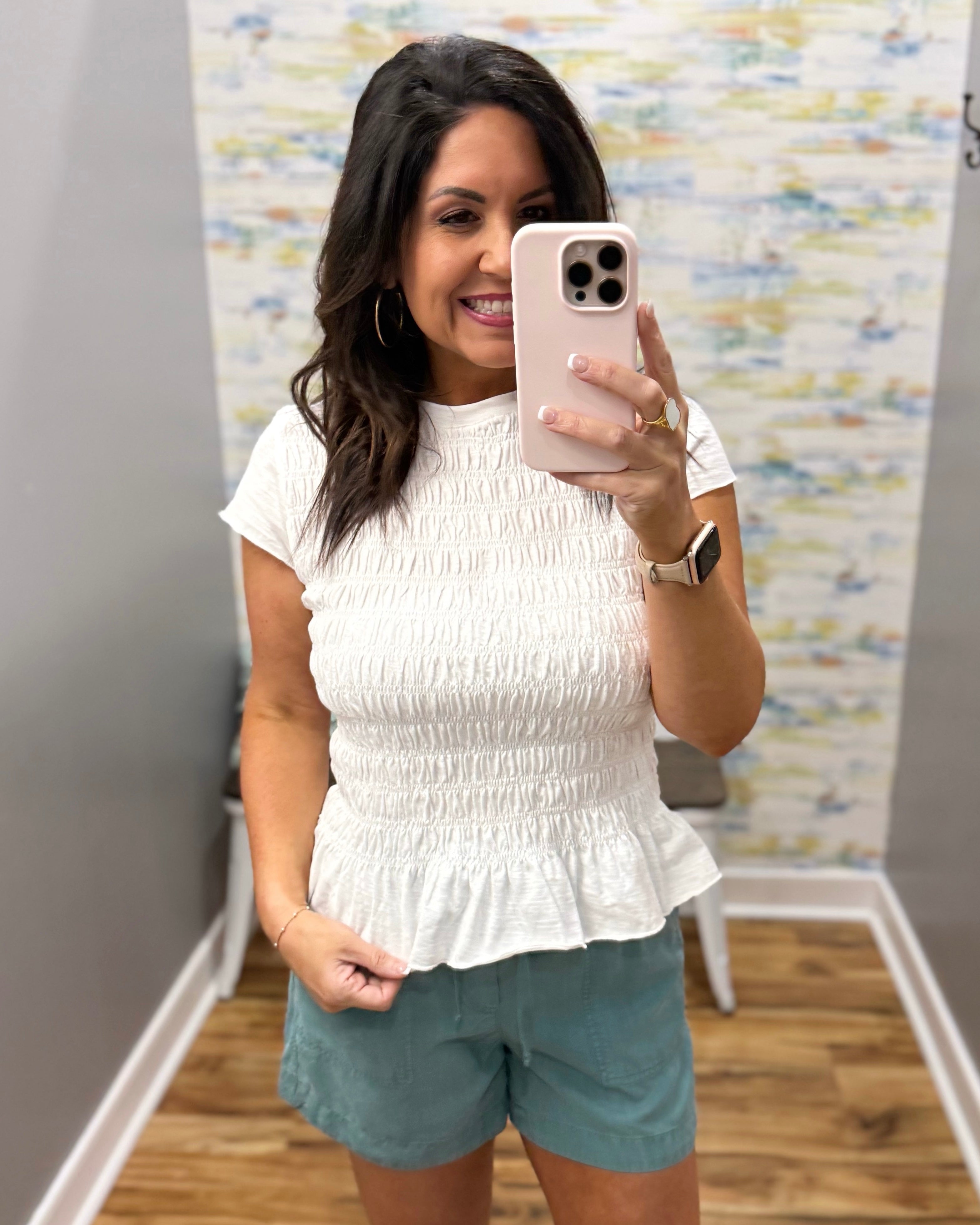 Woman taking a mirror selfie wearing a white top and green shorts in a room with colorful wall art.