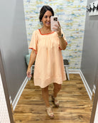 Woman taking a mirror selfie wearing a peach dress with red trim in a room with patterned wallpaper.