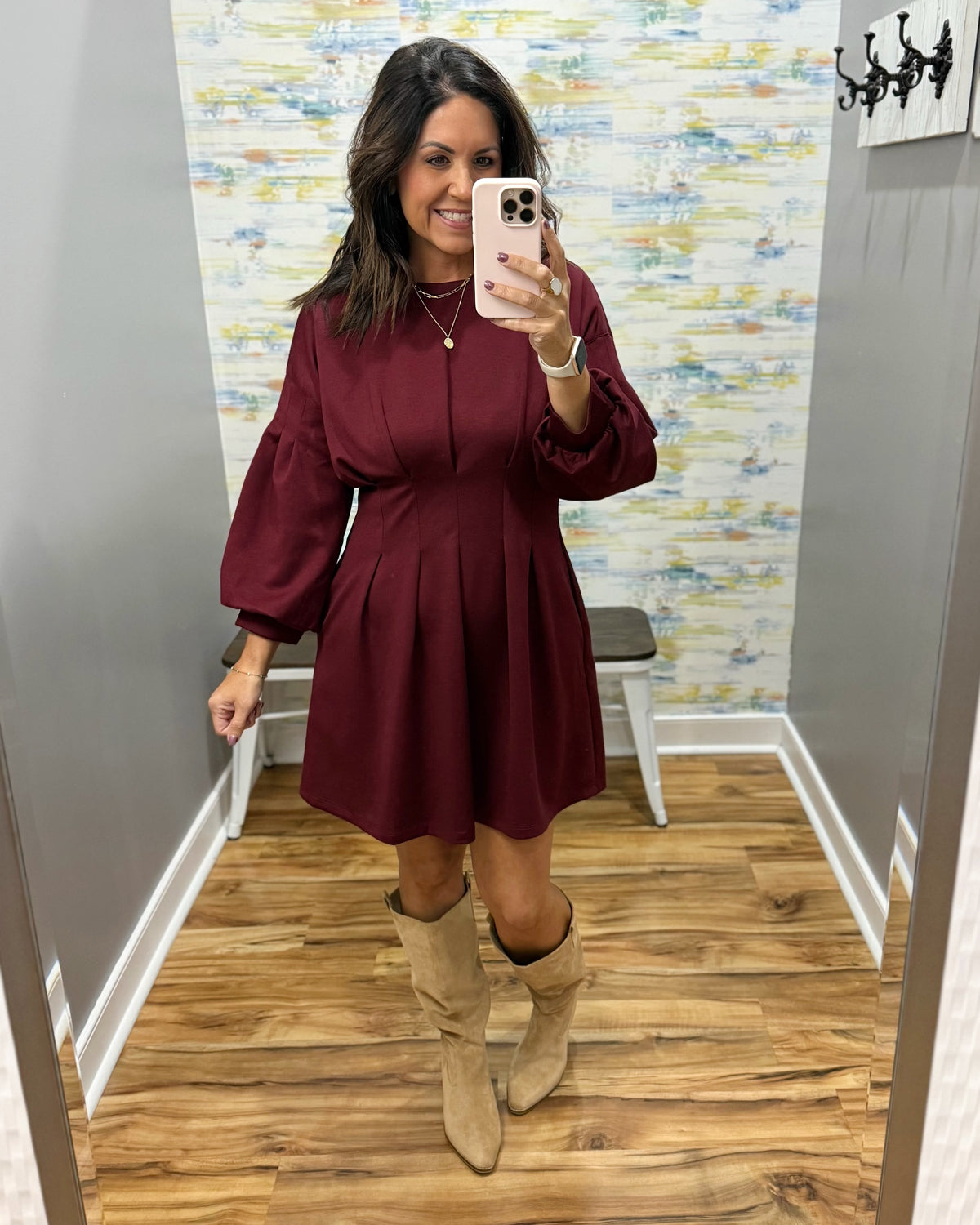 A woman takes a mirror selfie wearing a burgundy long-sleeve mini dress with puff sleeves and beige knee-high boots in a fitting room with wood floors and patterned wallpaper.