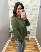 A woman takes a mirror selfie wearing an olive green long-sleeve crewneck sweater and light blue jeans in a fitting room with a wood floor and abstract wall art.