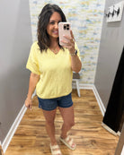 Woman taking a mirror selfie wearing a yellow t-shirt and denim shorts in a room with a colorful wall.
