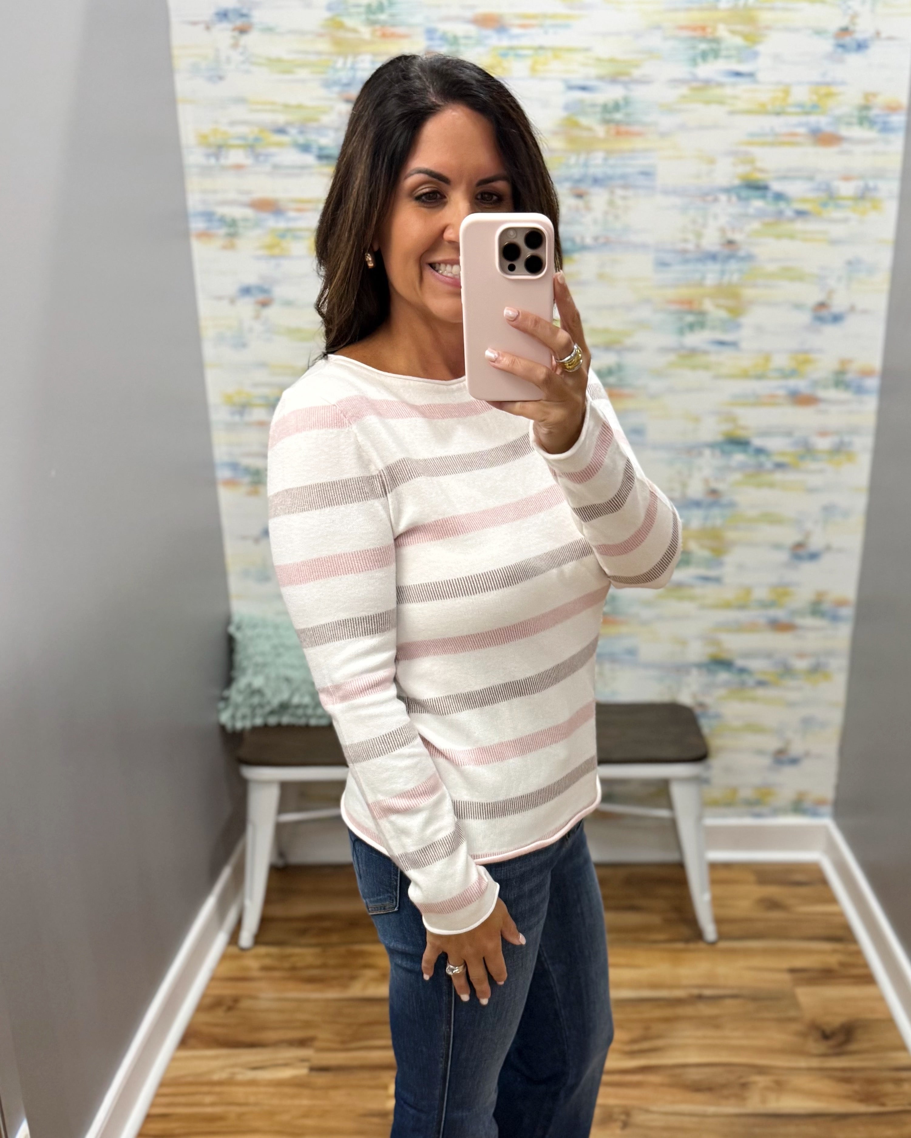 A woman takes a mirror selfie wearing a long-sleeve knit sweater with horizontal stripes in white, light pink, and taupe, paired with dark blue jeans.