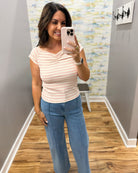 A woman takes a mirror selfie wearing a light peach and white striped short-sleeve top with blue jeans in a fitting room with wood floors and gray walls.
