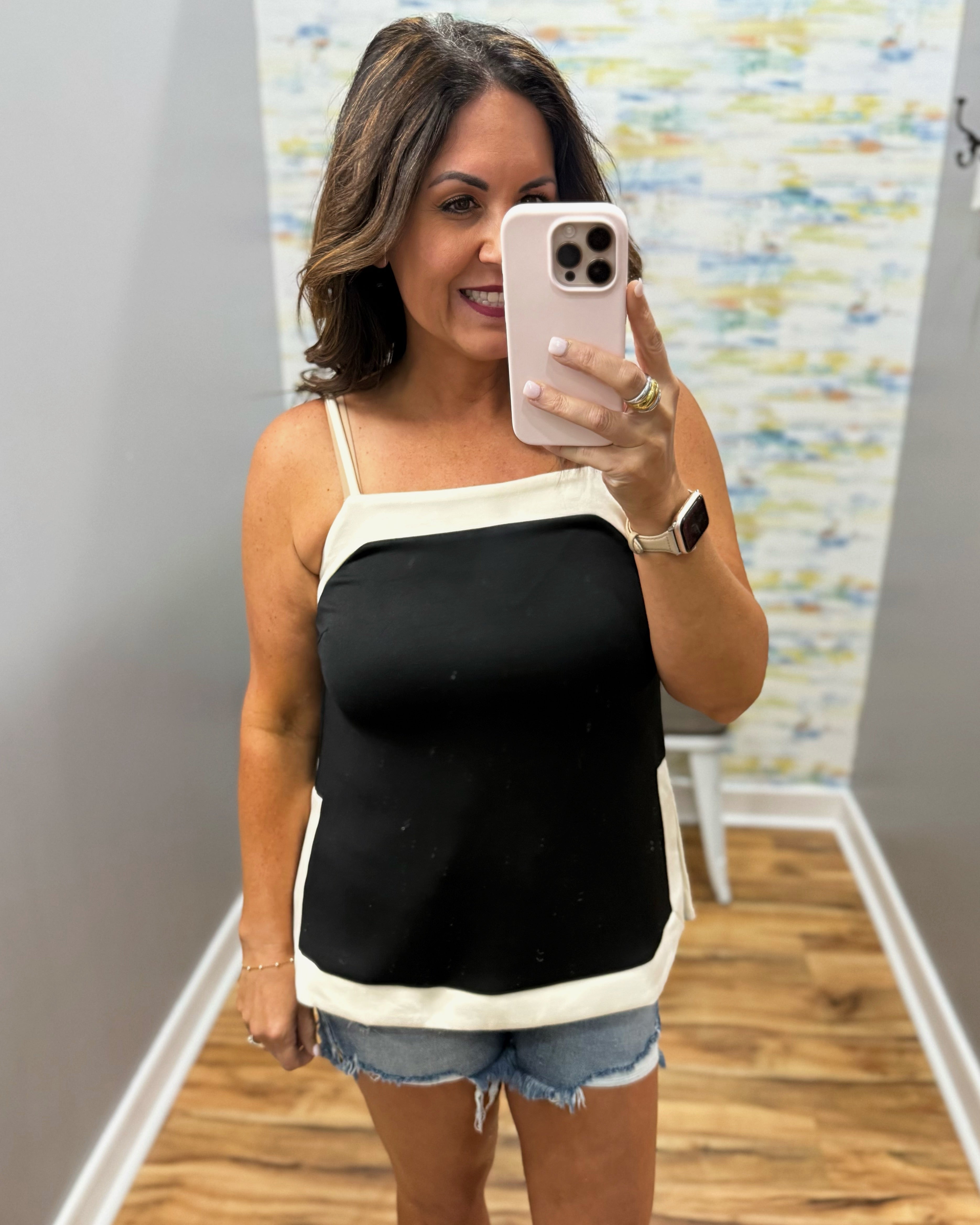 Woman taking a mirror selfie wearing a black and white tank top and denim shorts.