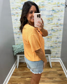 Woman taking a mirror selfie wearing a yellow top and denim shorts in a room with a colorful wall.