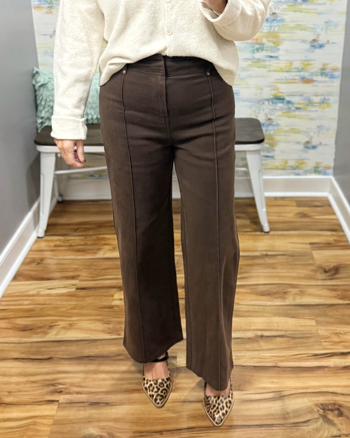 A person wearing dark brown wide-leg trousers with a cream knit top and leopard-print pointed flats, standing on a wood floor in an indoor room with a bench and abstract wall art.
