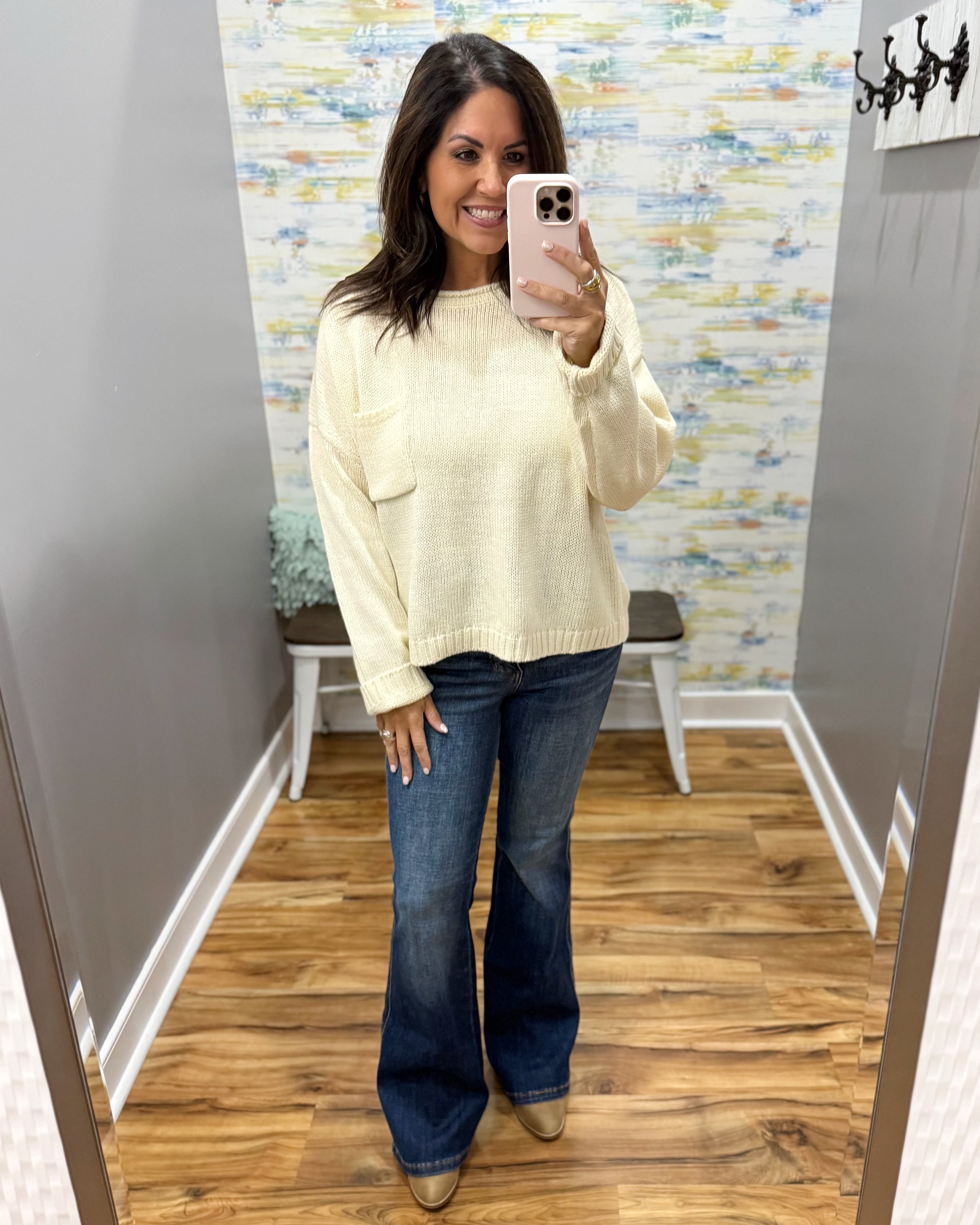 A woman takes a mirror selfie in a fitting room wearing a cream-colored knit sweater and dark blue flare jeans.