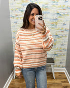 A woman takes a mirror selfie wearing a fuzzy, striped knit sweater in peach, orange, and cream, paired with blue jeans.