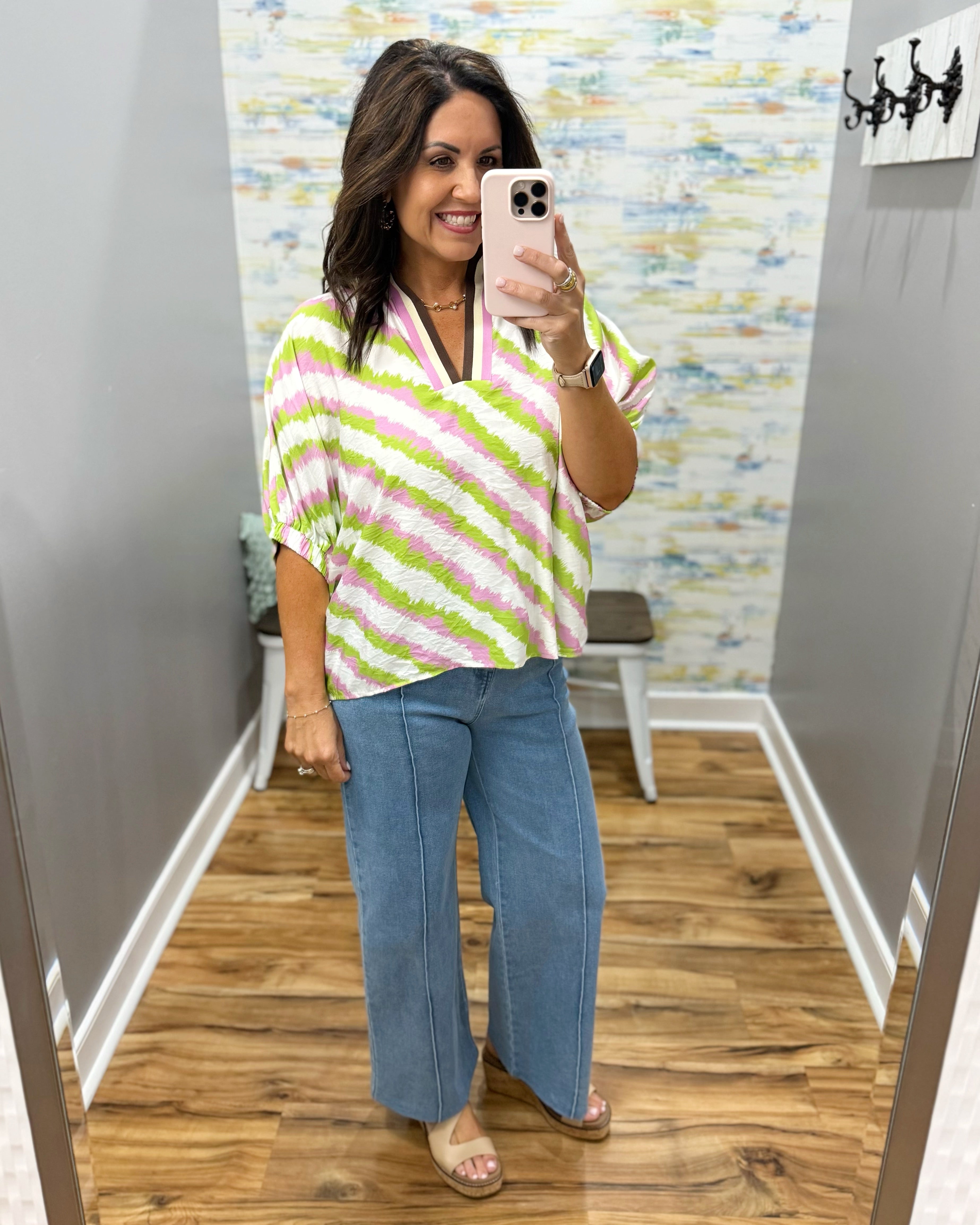 Woman taking a mirror selfie wearing a colorful striped top and blue jeans in a room with patterned wallpaper.