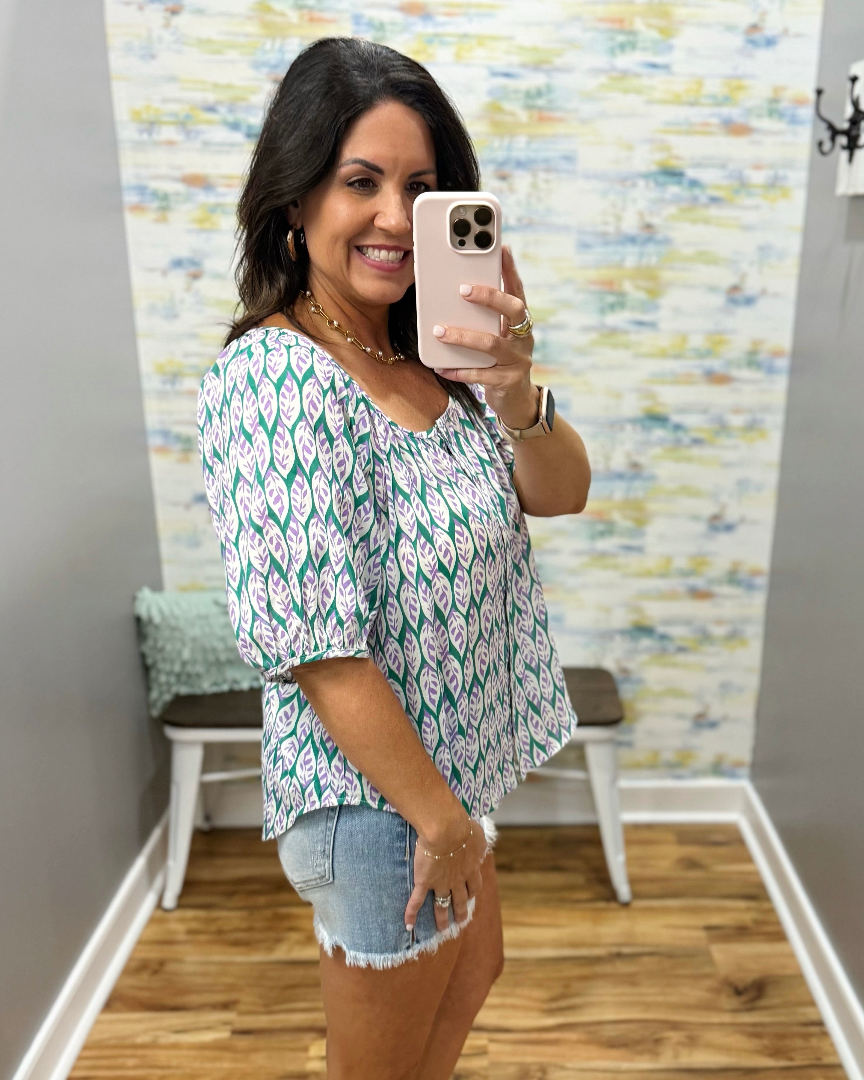 Woman taking a mirror selfie wearing a patterned top and denim shorts in a room with a textured wall.