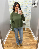 A woman takes a mirror selfie in a fitting room wearing an olive green long-sleeve crewneck sweater, blue jeans, and tan boots.