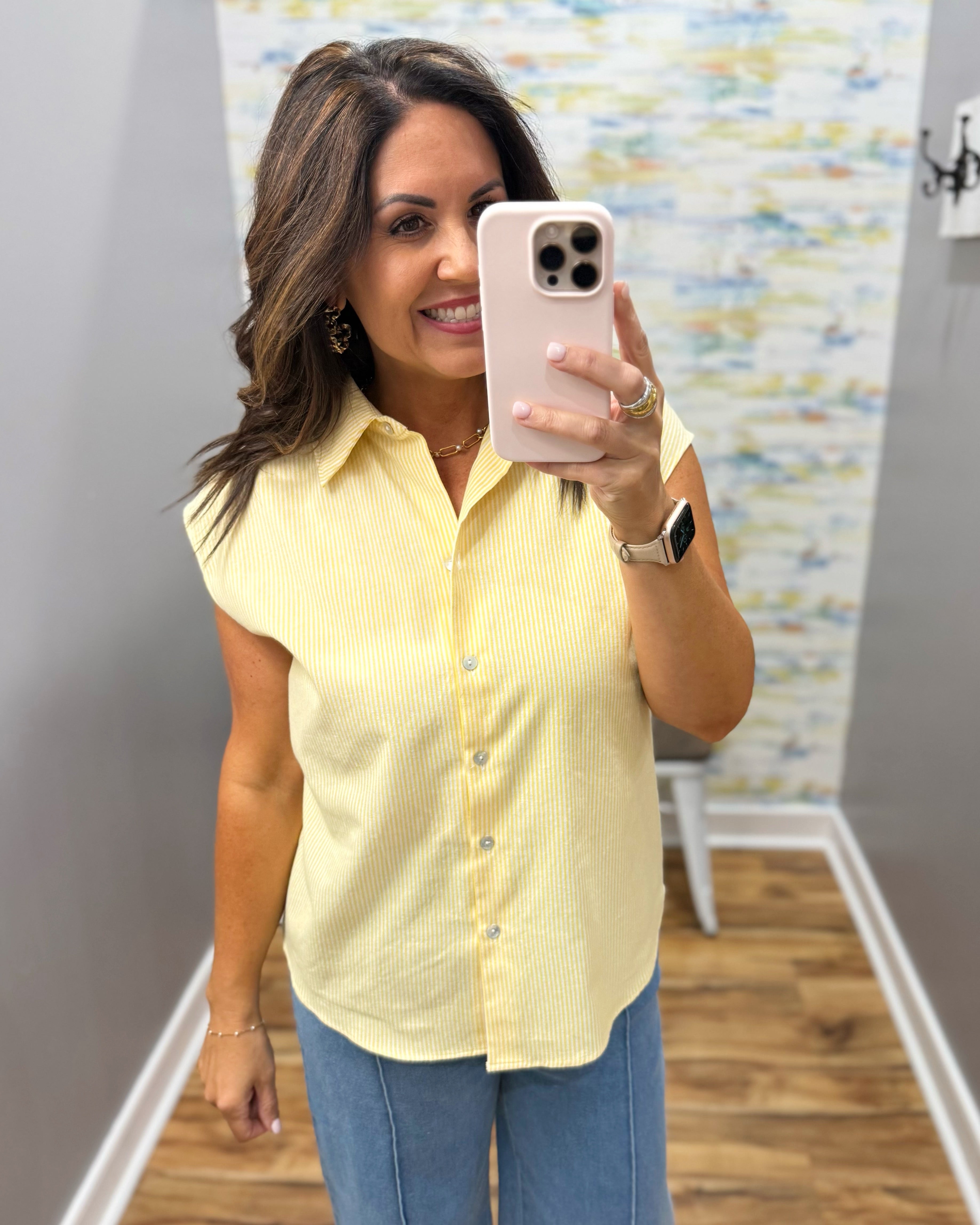 Woman taking a mirror selfie wearing a yellow shirt and blue jeans in a room with a patterned wall.