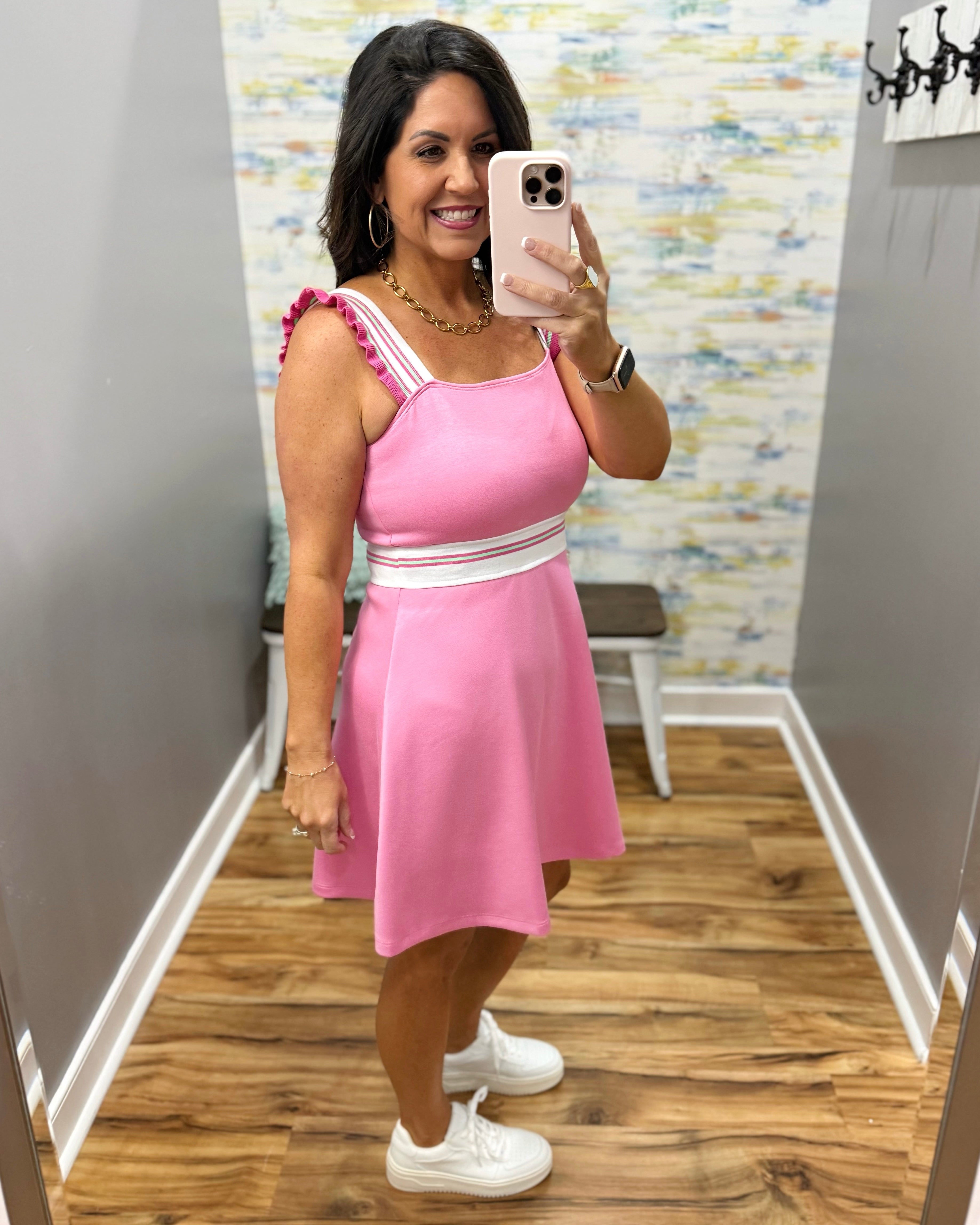 Woman taking a mirror selfie wearing a pink dress with white accents in a room with a patterned wall.