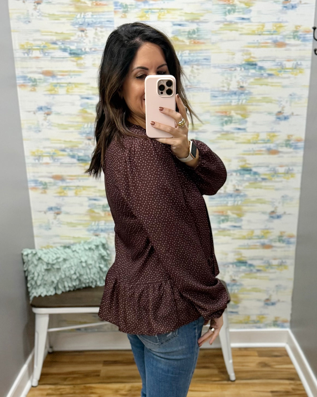 A woman takes a mirror selfie in a fitting room wearing a dark brown long-sleeve blouse with a tiny white polka dot pattern and blue jeans.