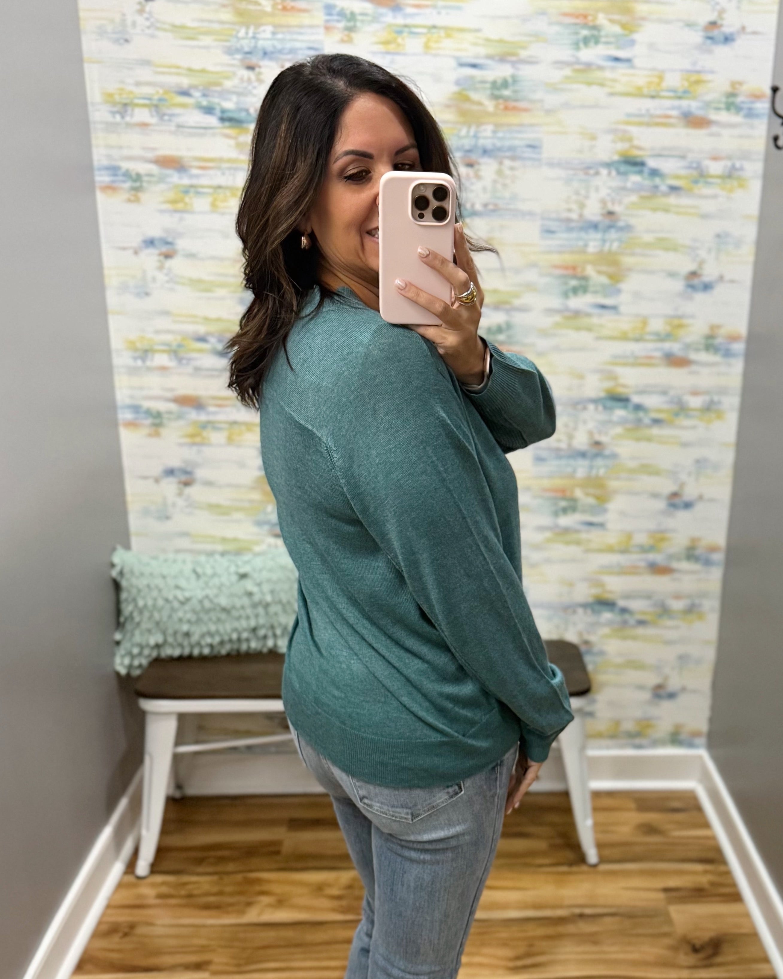 A woman takes a mirror selfie wearing a teal green long-sleeve knit top and light-wash blue jeans in a home setting with a patterned wallpaper background.