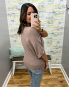 A woman takes a mirror selfie in a fitting room wearing a loose, short-sleeve knit top in a muted taupe-brown color, paired with light-wash blue jeans.