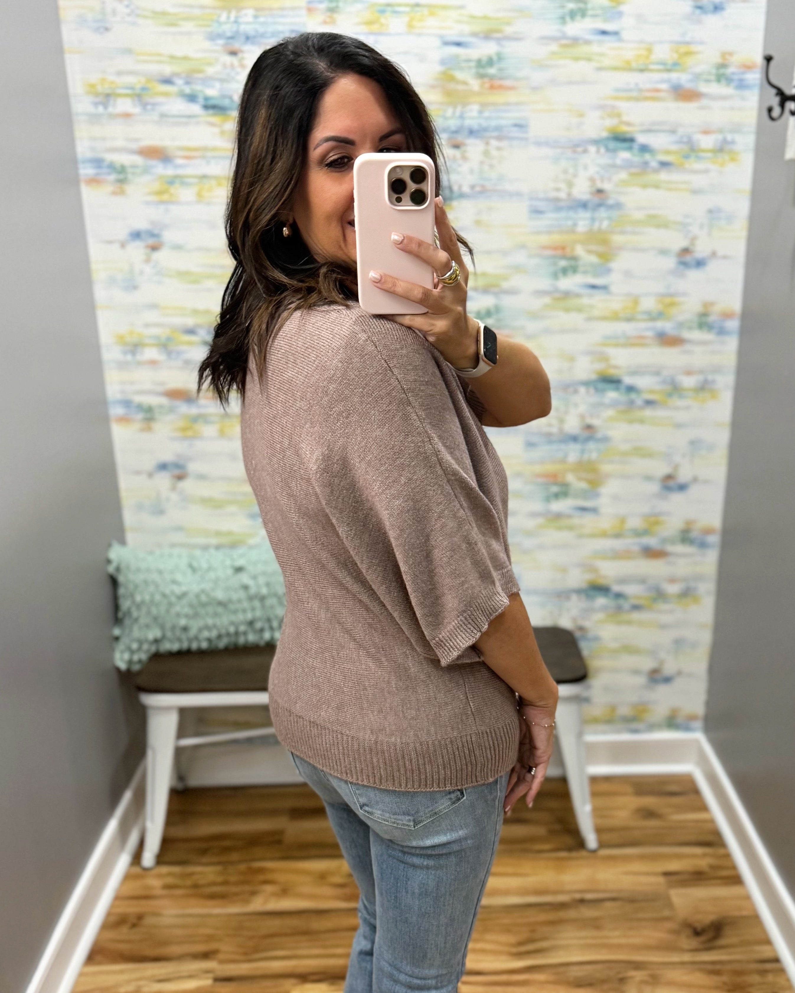 A woman takes a mirror selfie in a fitting room wearing a loose, short-sleeve knit top in a muted taupe-brown color, paired with light-wash blue jeans.