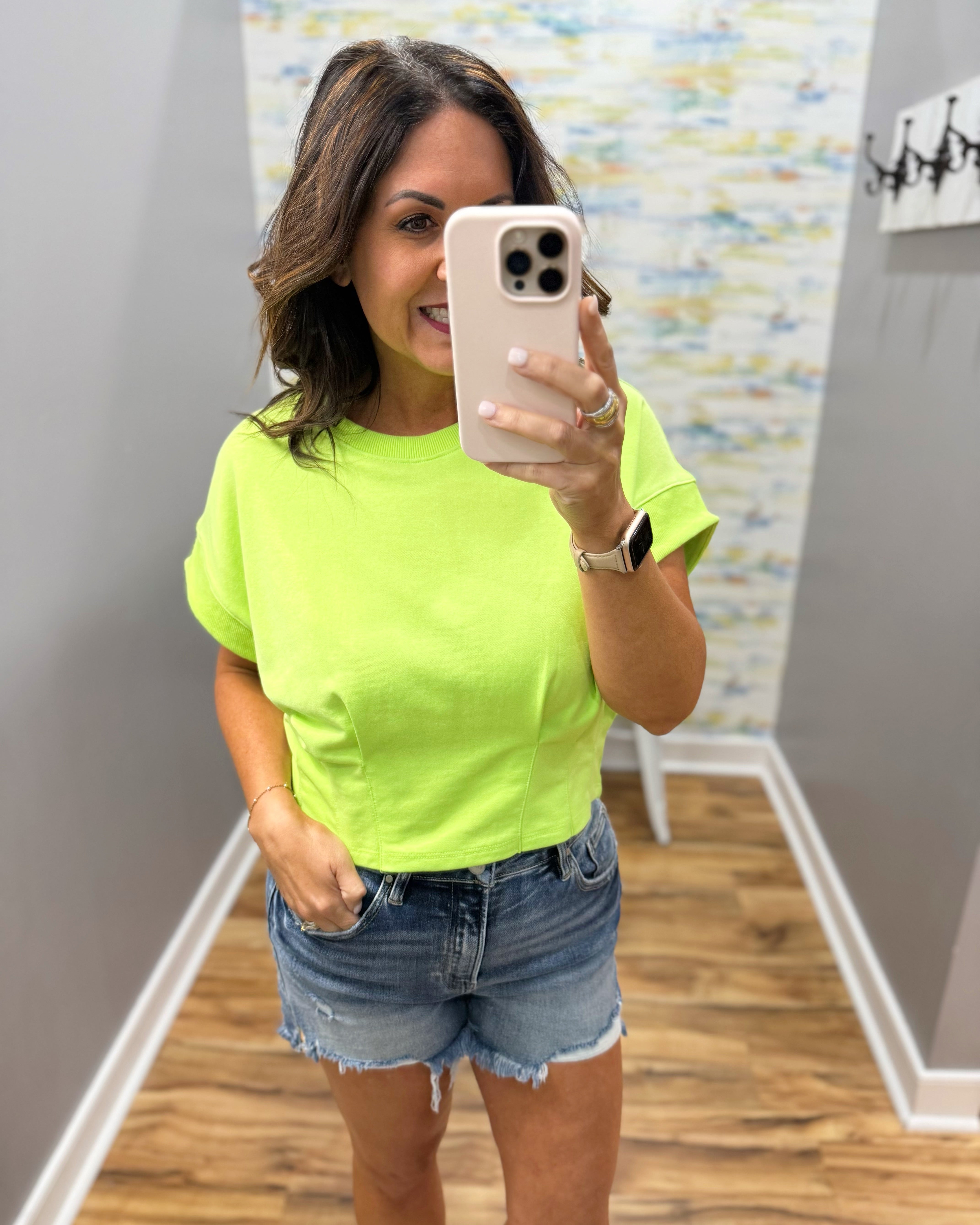 Woman taking a mirror selfie wearing a bright green t-shirt and denim shorts.