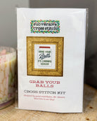 A packaged cross stitch kit labeled 'subversive cross stitch' with the design text 'Grab Your Balls / It's Canning Season,' shown in clear plastic wrapping on a tabletop next to a candle.