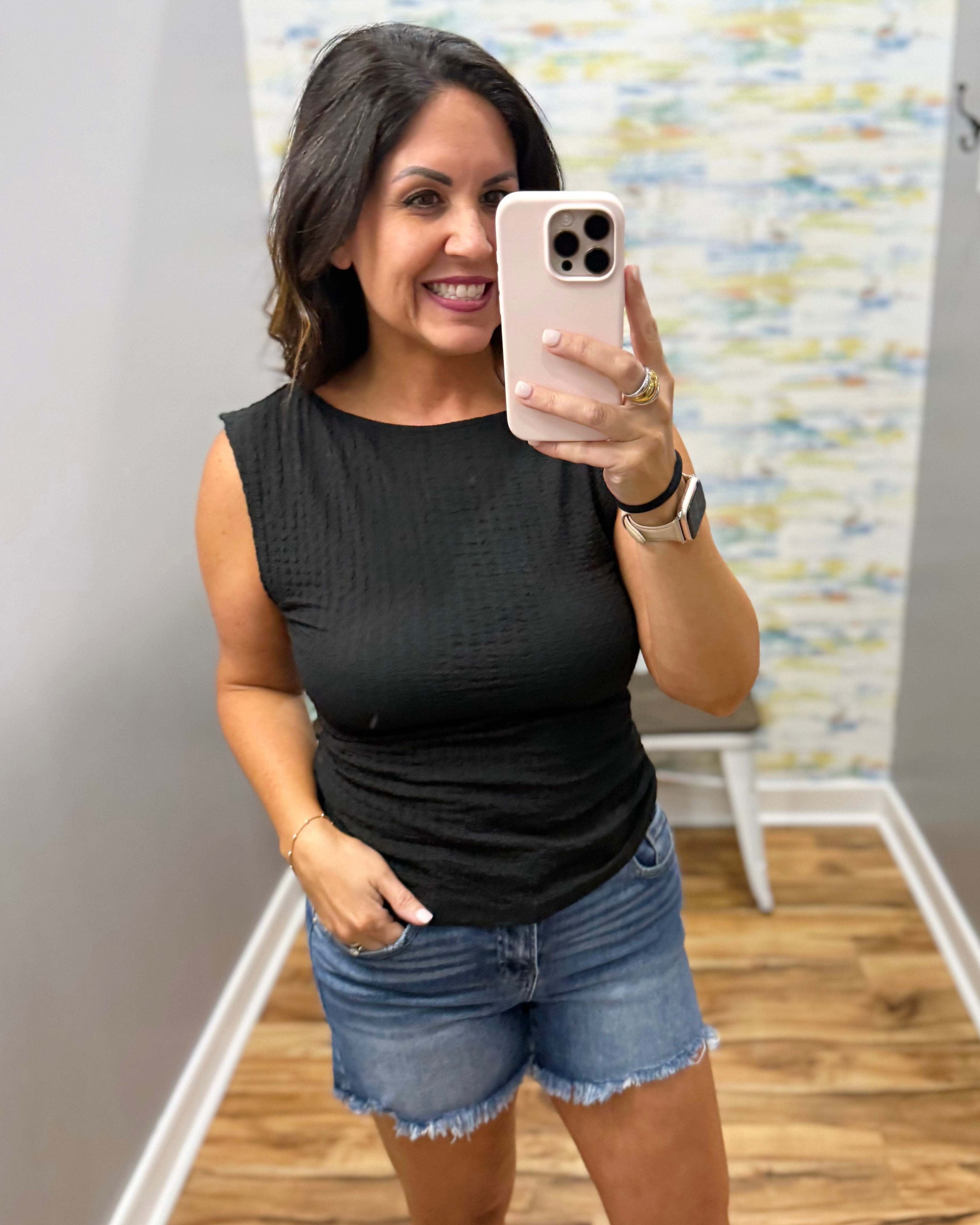 Woman taking a mirror selfie wearing a black sleeveless top and denim shorts.
