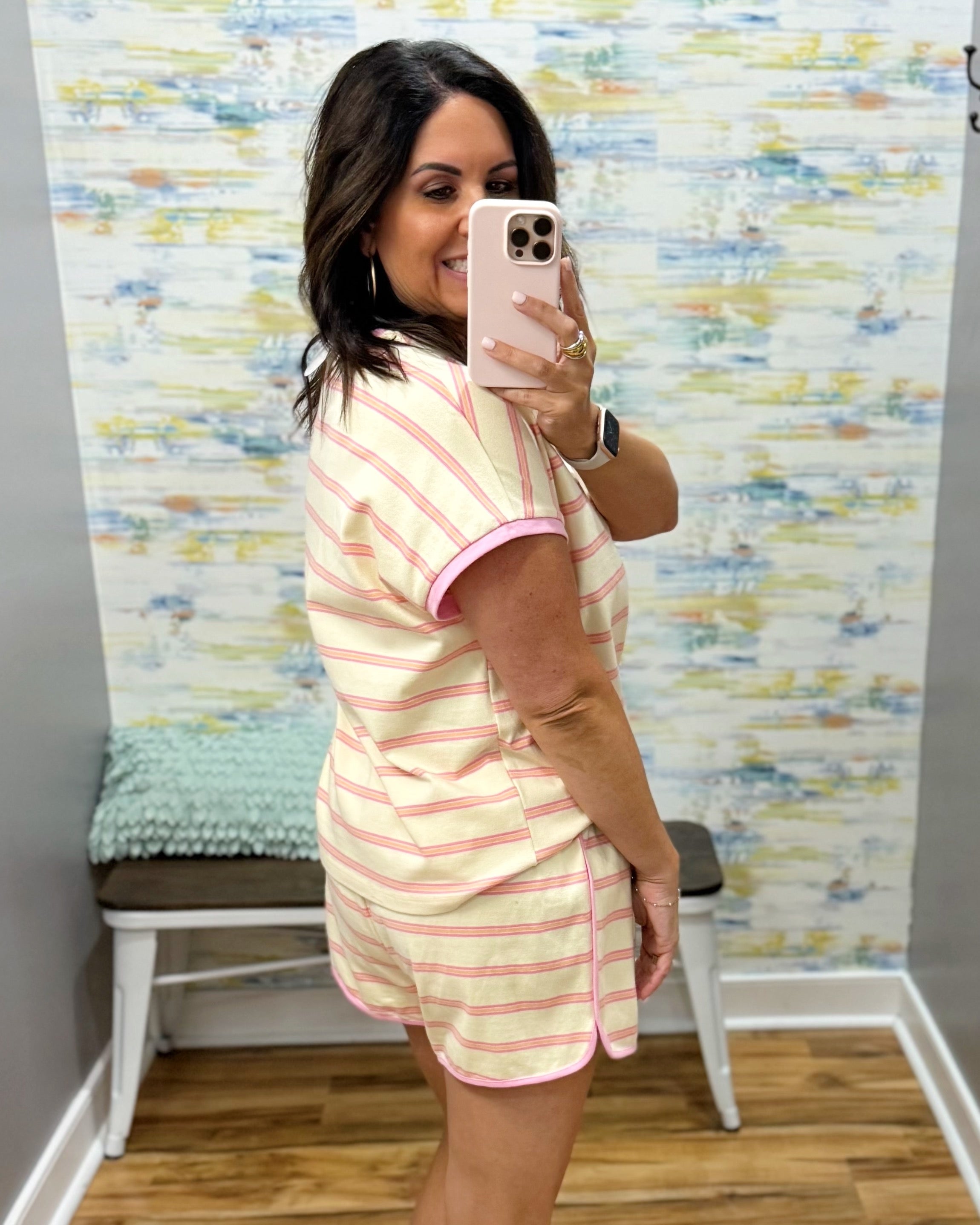 A woman takes a mirror selfie wearing a matching two-piece pajama set with light pink and white horizontal stripes. The set includes a short-sleeve crewneck t-shirt and matching shorts, shown in a home or boutique fitting room environment.