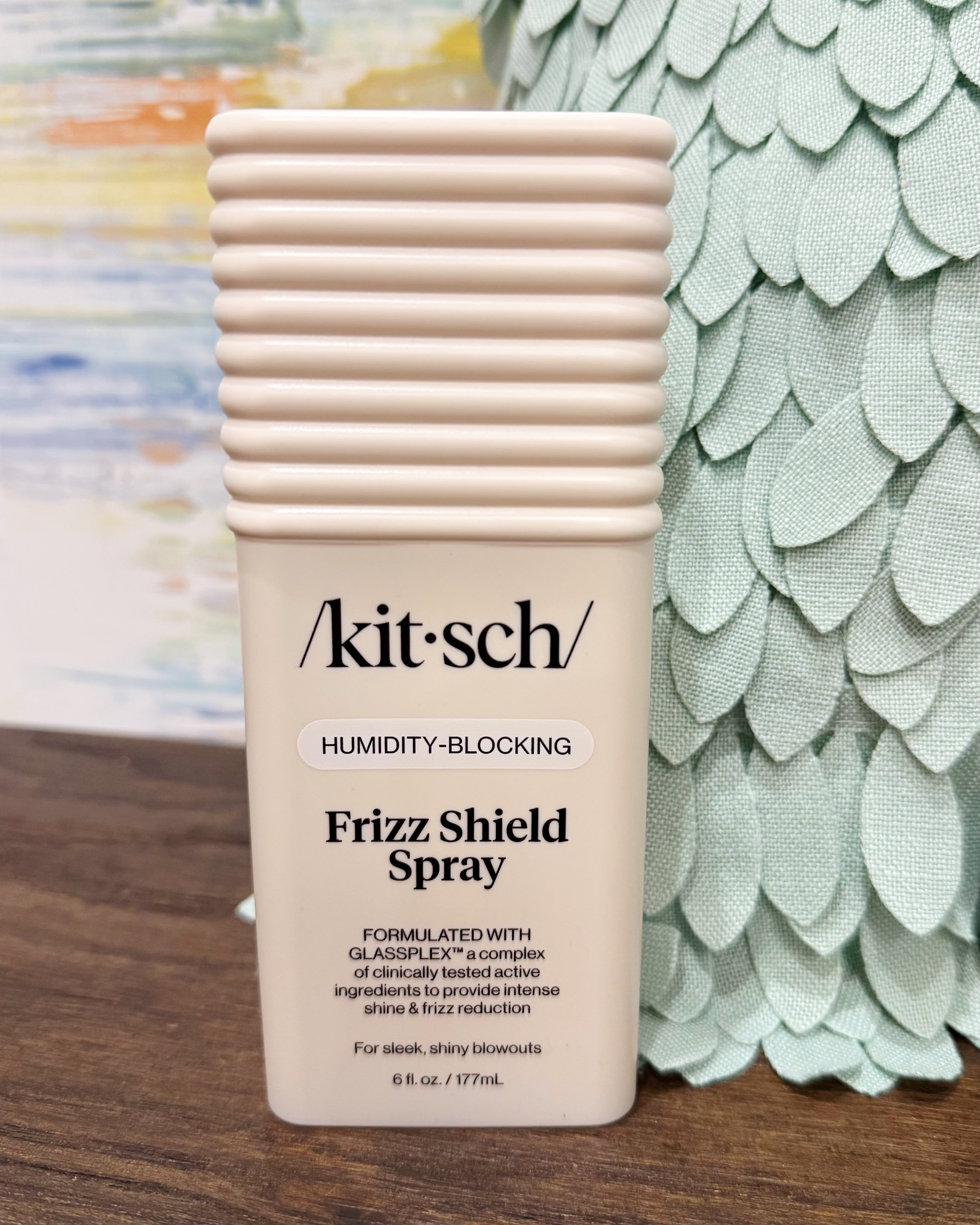 Kit-sch Frizz Shield Spray bottle in a pale peach-beige color, standing on a wooden surface with a decorative green textured vase in the background.