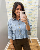 A woman taking a mirror selfie wearing a light-wash blue denim shirt with rolled sleeves, gold-tone buttons, and a ruffled hem, paired with brown pants.