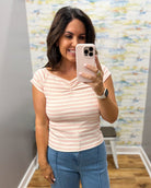 A woman taking a mirror selfie in a fitting room wearing a light pink and white striped short-sleeve top with a ruched V-neckline, paired with blue jeans.
