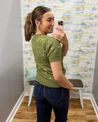 A woman takes a mirror selfie wearing an olive green short-sleeve knit sweater with small white embroidered dots, paired with dark blue jeans in a fitting room.