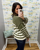 A woman takes a mirror selfie in a fitting room wearing an olive green and cream striped knit sweater, blue jeans, and holding a light pink smartphone.