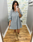 A woman takes a mirror selfie wearing a blue and white vertically striped button-down shirt dress with three-quarter sleeves, paired with beige suede boots, in a fitting room with wood floors and abstract wallpaper.