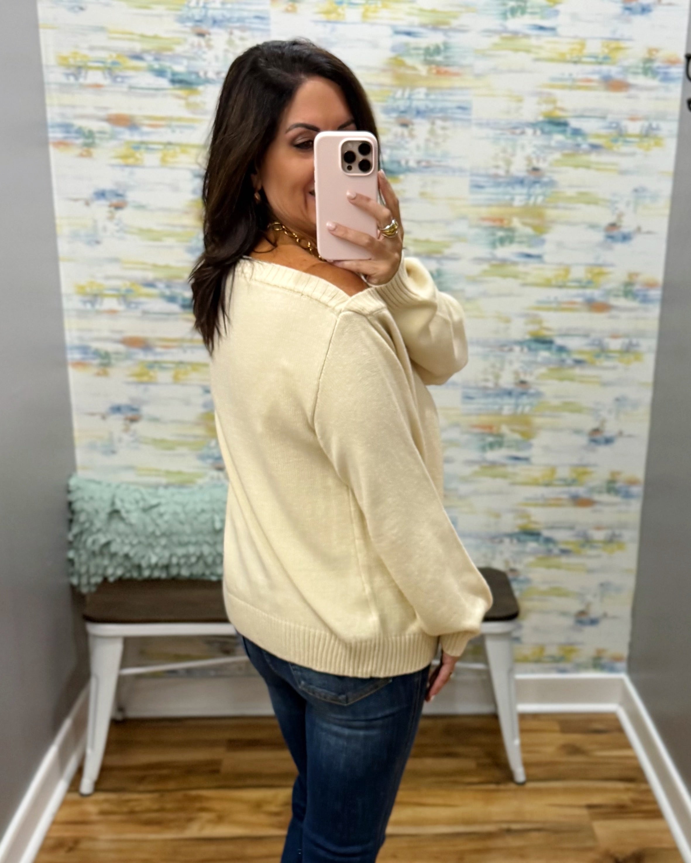 A woman takes a mirror selfie in a boutique showing a cream-colored knit sweater with blue jeans, standing in front of a patterned wallpaper background.
