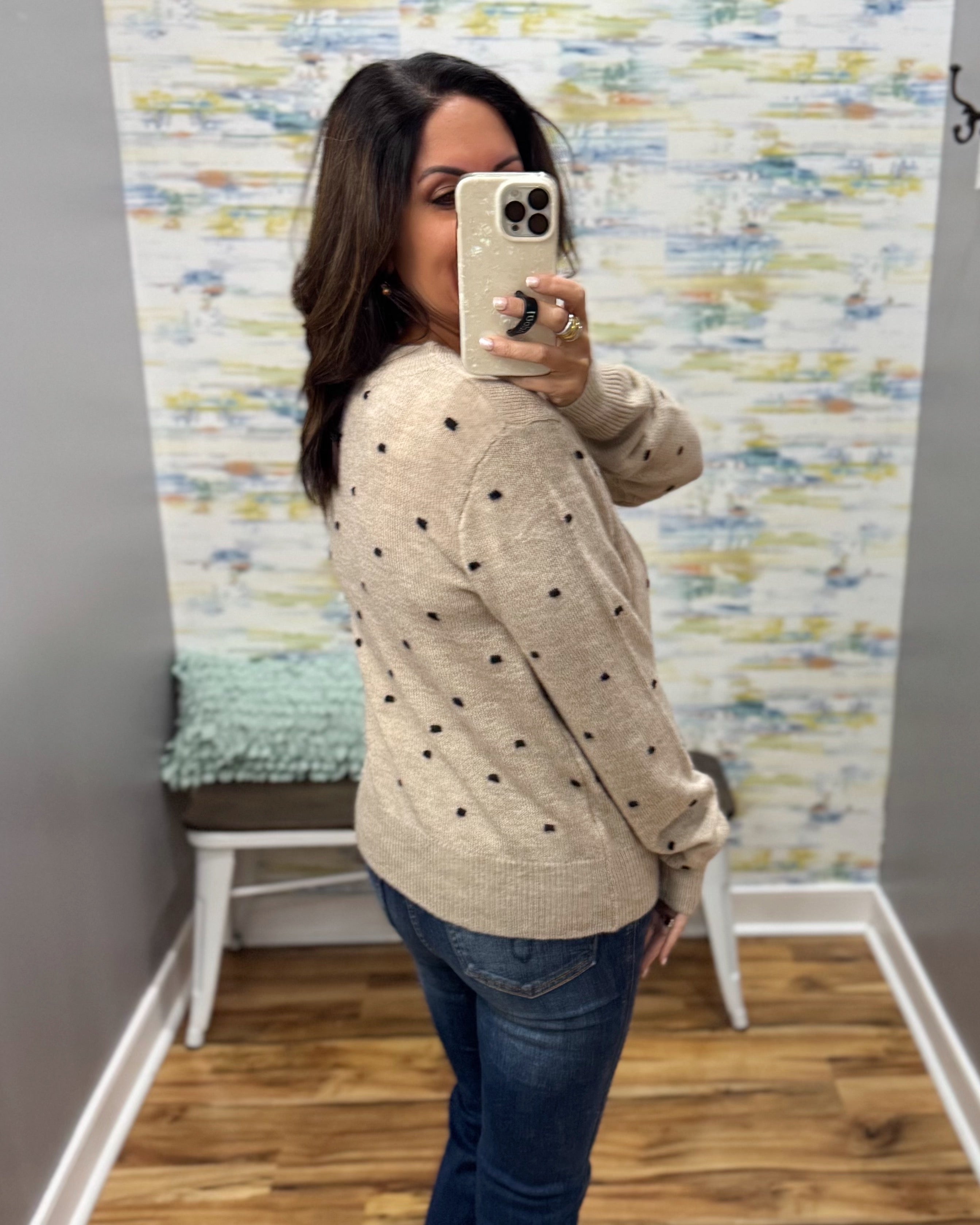 A woman takes a mirror selfie in a fitting room wearing a beige knit sweater with black polka dots and blue jeans.