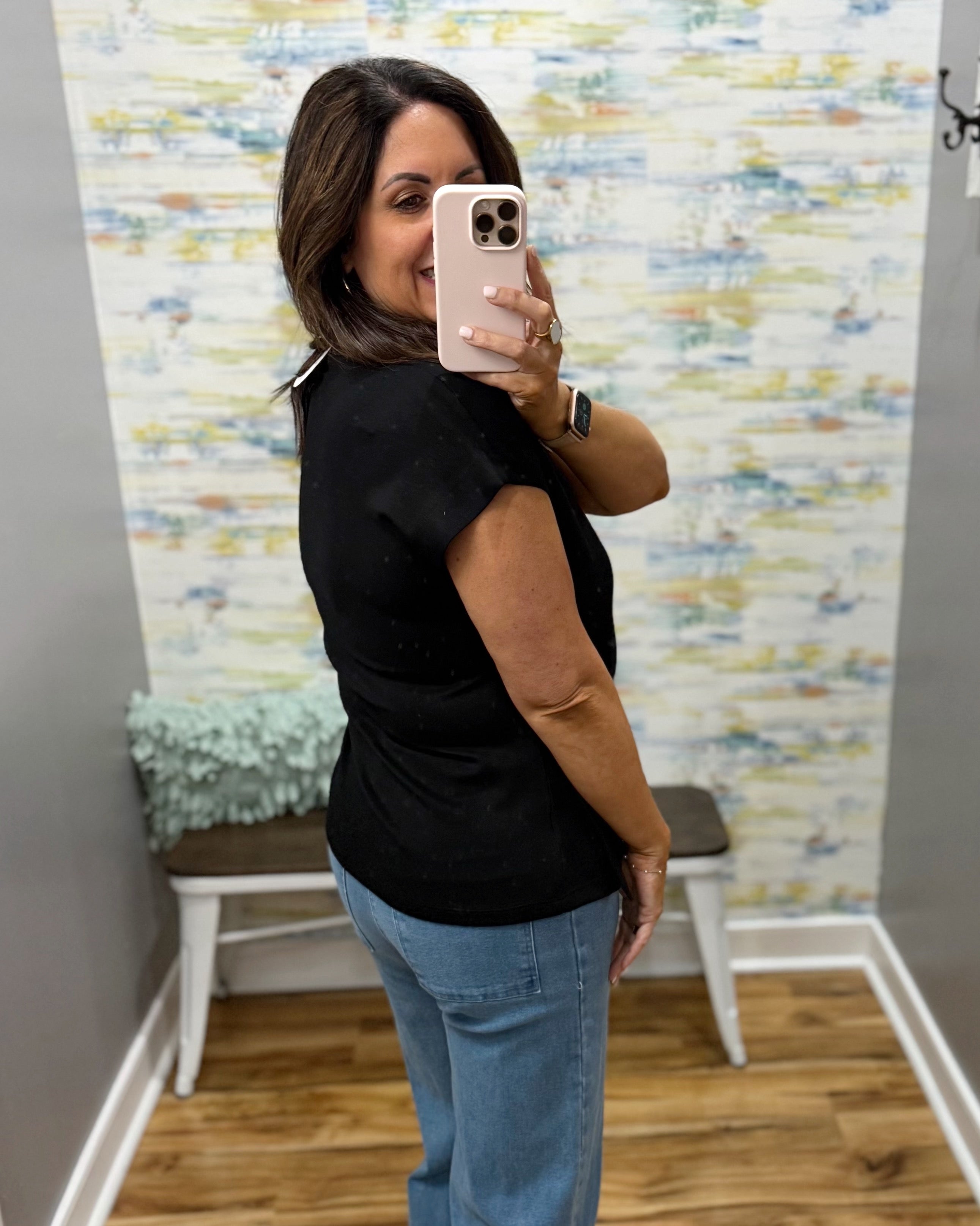 Woman taking a mirror selfie wearing a black top and blue jeans in a room with colorful wall art.
