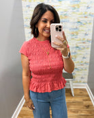 A woman takes a mirror selfie wearing a coral pink short-sleeve smocked peplum top and blue jeans in a fitting room.