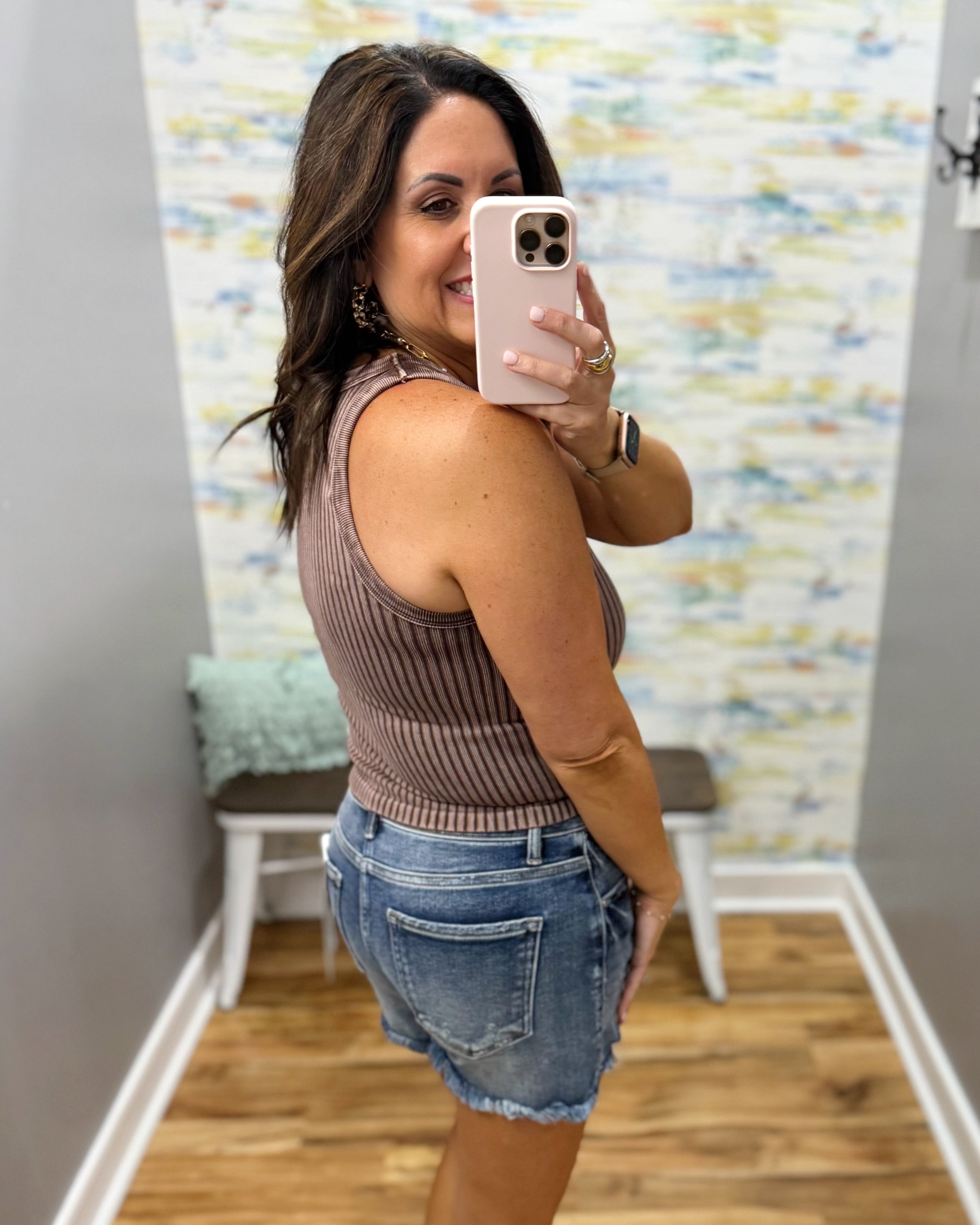 Woman taking a mirror selfie wearing a brown ribbed tank top and denim shorts in a room with a colorful wall.