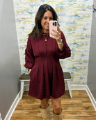 A woman takes a mirror selfie wearing a long-sleeve, button-front, pleated mini dress in deep burgundy, paired with beige boots. The background shows a gray wall, a colorful abstract-patterned wallpaper, a white bench, and a wood floor.