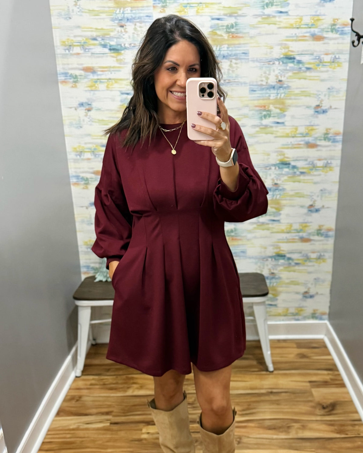 A woman takes a mirror selfie wearing a long-sleeve, button-front, pleated mini dress in deep burgundy, paired with beige boots. The background shows a gray wall, a colorful abstract-patterned wallpaper, a white bench, and a wood floor.
