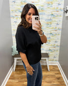 A woman takes a mirror selfie in a fitting room wearing a black short-sleeve knit sweater and blue jeans.