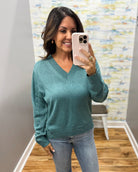 A woman takes a mirror selfie wearing a teal green long-sleeve V-neck knit sweater and light-wash blue jeans in an indoor room with wood floors and a patterned wall.