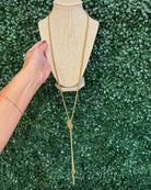 Chansuttpearls gold lariat-style necklace with a knotted center detail is displayed on a beige fabric mannequin bust. A person's hand with dark nail polish is visible on the left side holding the display bust, set against a green artificial foliage background.